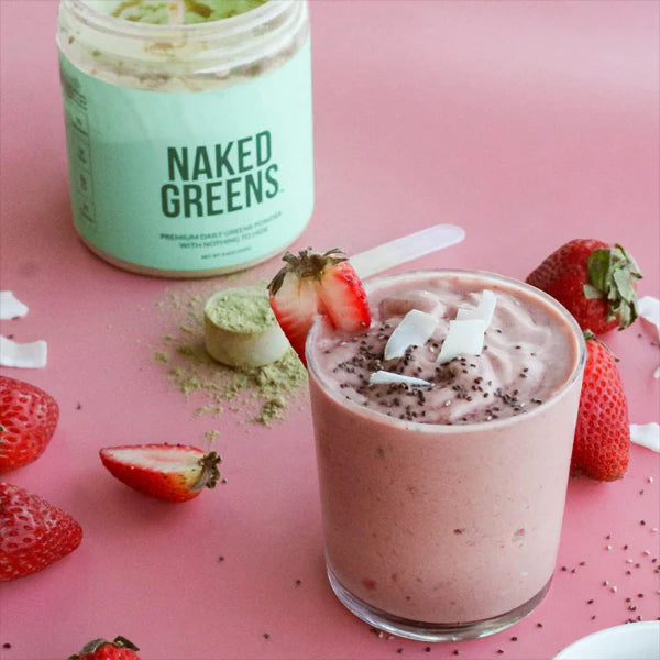 A glass of pink smoothie with chia seeds, coconut flakes, and a strawberry garnish sits in front of whole strawberries and a jar of Naked Nutrition’s Naked Super Greens Powder Organic Greens Supplement 8.6oz.